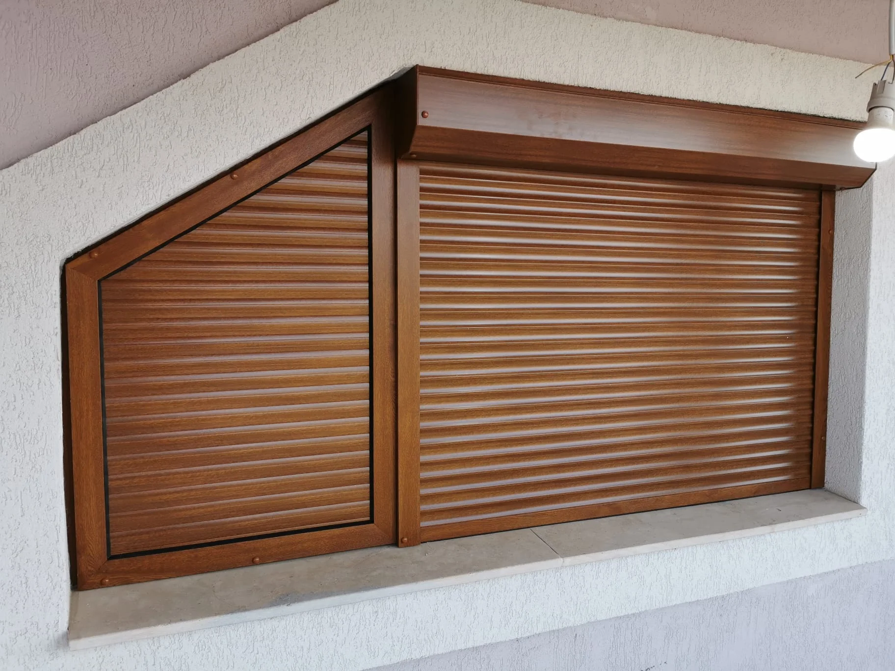 Două ferestre alăturate, una cu ramă în unghi și una dreptunghiulară, cu rulouri exterioare din aluminiu în nuanță stejar auriu, montate pe fațada unei case